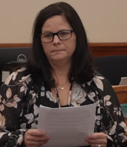 A photo of Rhonda Jewell, a woman with dark hair wearing a blouse with a floral pattern. She is holding documents in her hand.